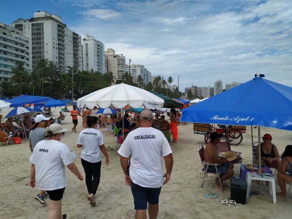 De camisetas brancas, com a palavra fiscalização escrita atrás, agentes caminham entre banhistas na praia. No canto direito, pessoas estão sentadas debaixo de um guarda sol azul. Ao fundo há mais guarda sol e outros banhistas.