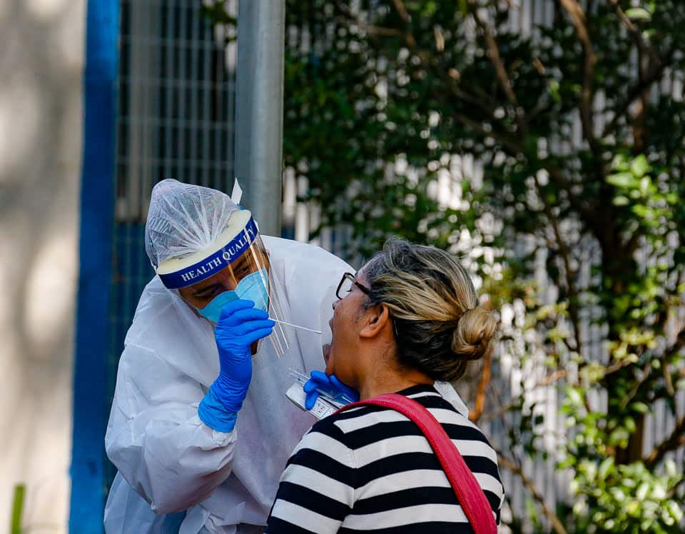 agente de saúde, usando equipamentos de proteção nas mãos e no rosto, coleta amostra de saliva de paciente mulher. Na foto, a mulher aparece com a boca aberta e a agente de saúde inserindo uma haste na boca para fazer a coleta.